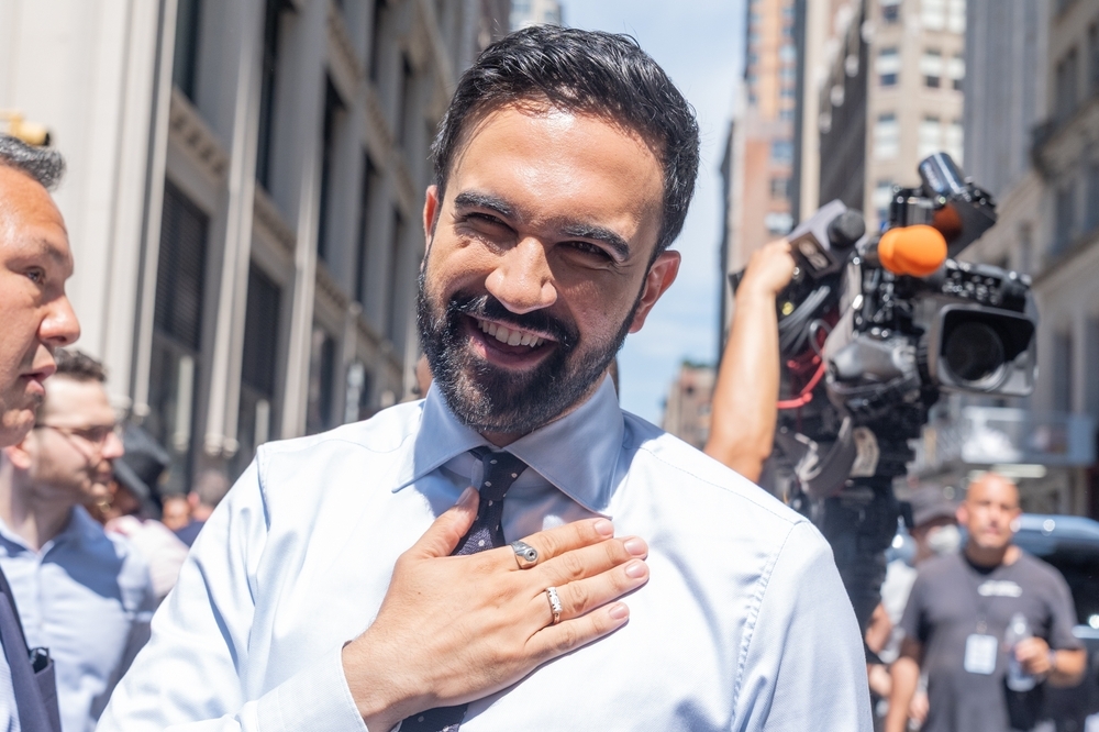 Zohran Mamdani souriant en public, main sur le plexus, entouré de caméras à New York, il est sorti victorieux de la primaire démocrate de 2025